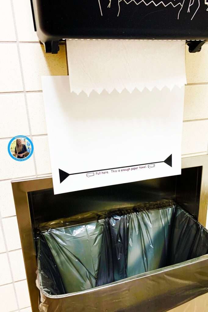 A poster under a paper towel dispenser in a school restroom that reads, "Pull here. This is enough paper towel."